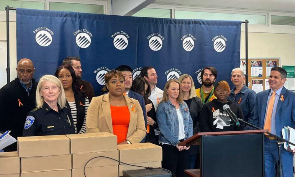 Group of diverse officials and community members at a press conference in front of a Pierce County backdrop, with stacked boxes on the table and a podium with microphones.