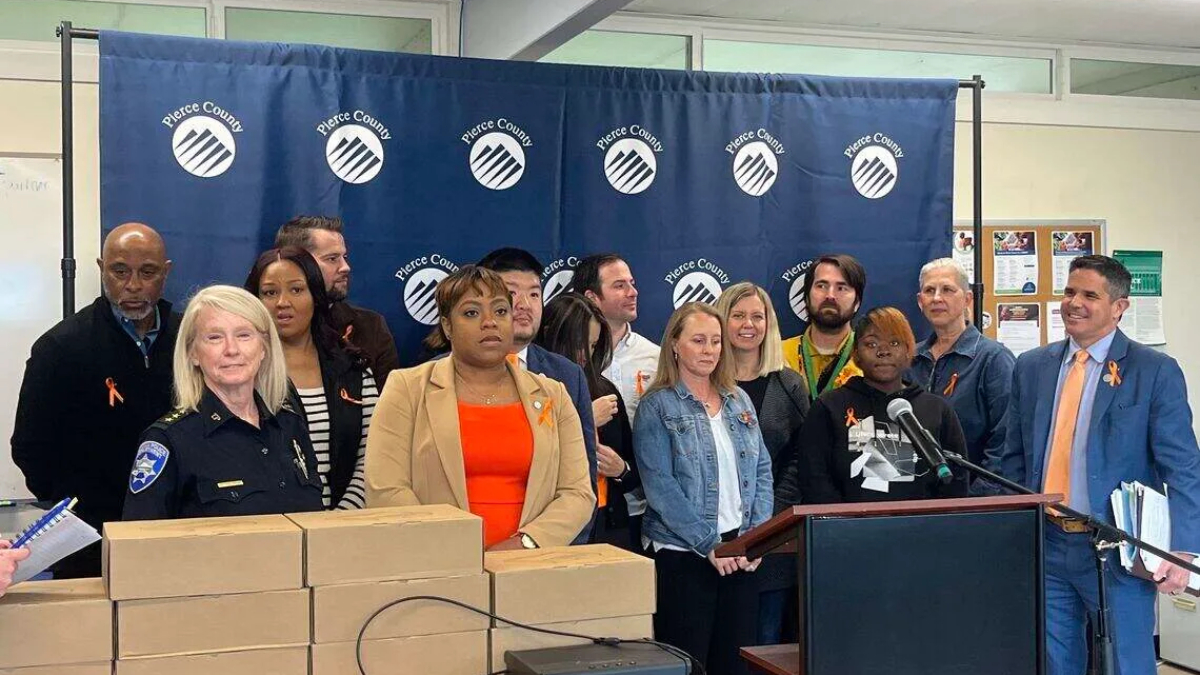Group of diverse officials and community members at a press conference in front of a Pierce County backdrop, with stacked boxes on the table and a podium with microphones.