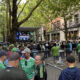 Crowd gathered along a city street watching a live concert on a stage with performers.