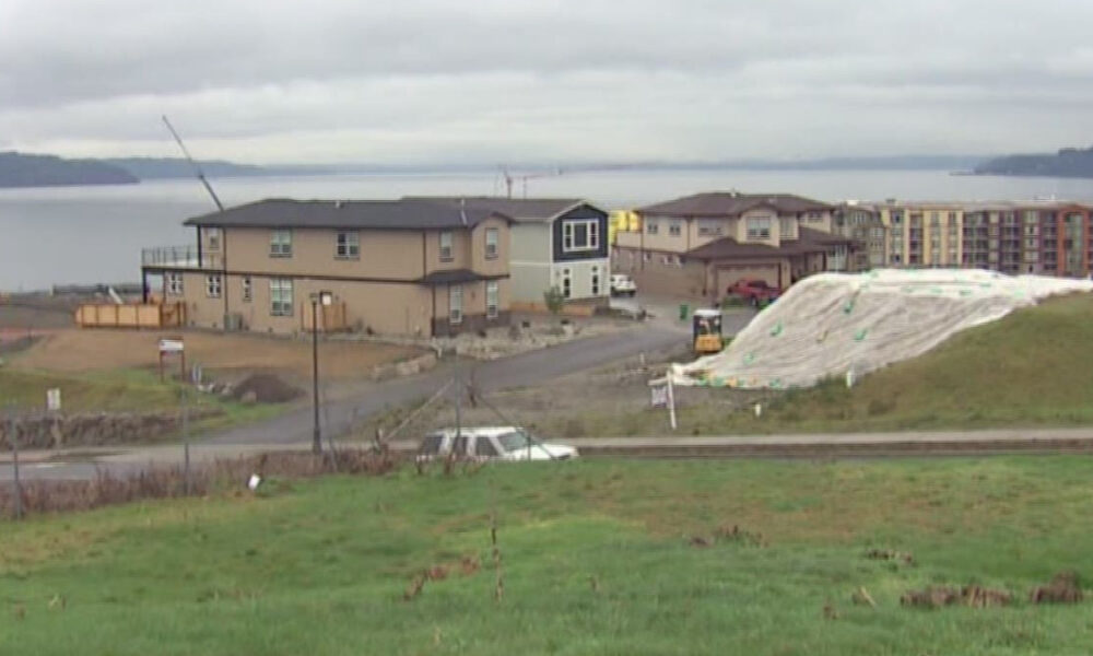 Residential construction by a lake: beige two-story houses, dirt road, parked car, and a large white tarp-covered slope in the foreground.
