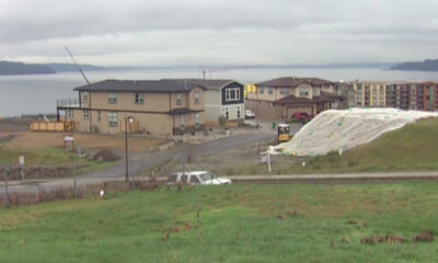 Residential construction by a lake: beige two-story houses, dirt road, parked car, and a large white tarp-covered slope in the foreground.