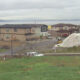 Residential construction by a lake: beige two-story houses, dirt road, parked car, and a large white tarp-covered slope in the foreground.