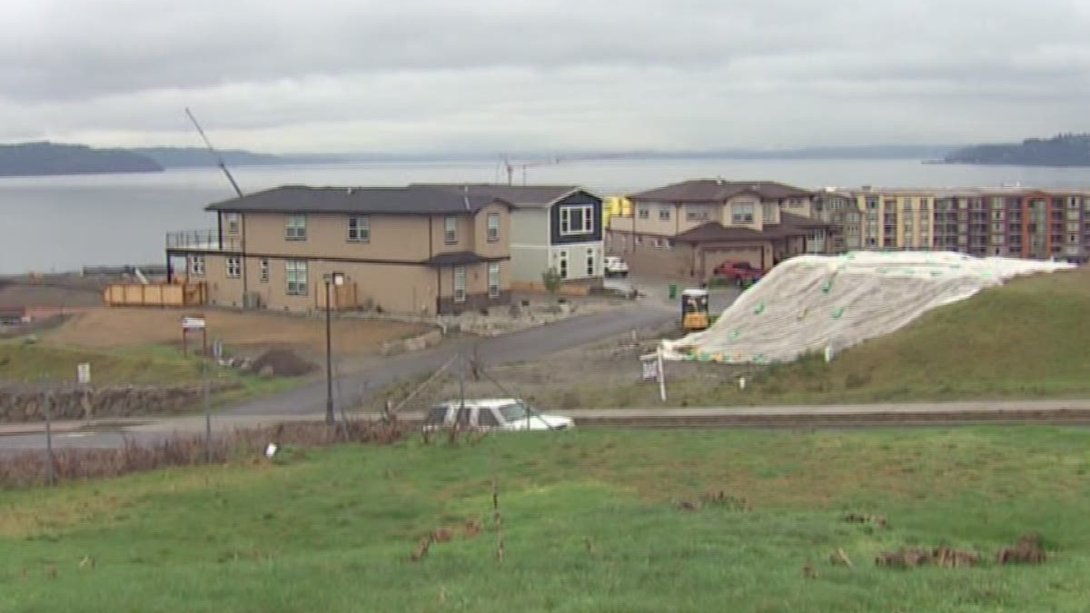 Residential construction by a lake: beige two-story houses, dirt road, parked car, and a large white tarp-covered slope in the foreground.