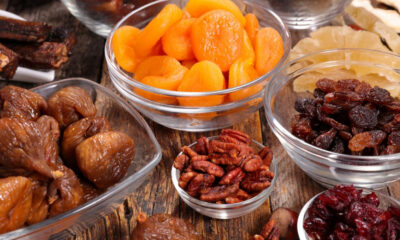 Assorted dried fruits in glass bowls (apricots, raisins, dates) arranged on a rustic wooden surface.