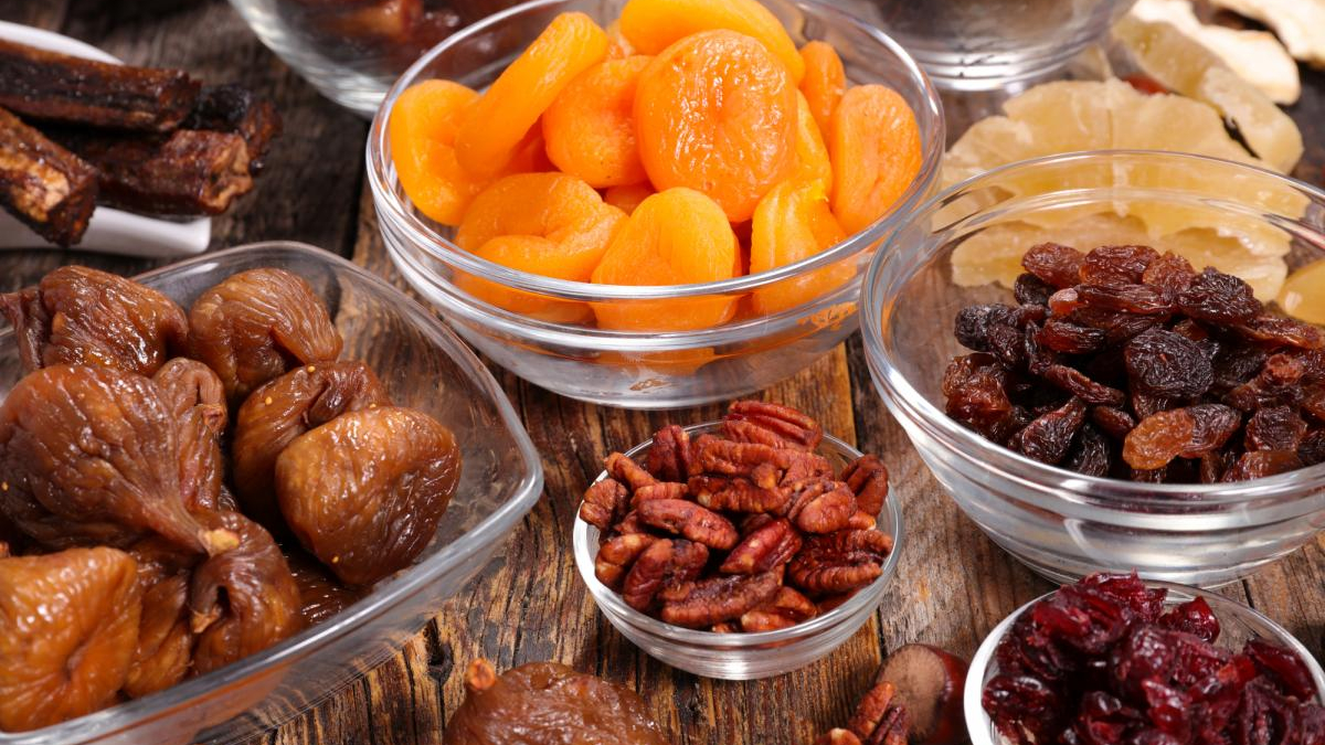 Assorted dried fruits in glass bowls (apricots, raisins, dates) arranged on a rustic wooden surface.
