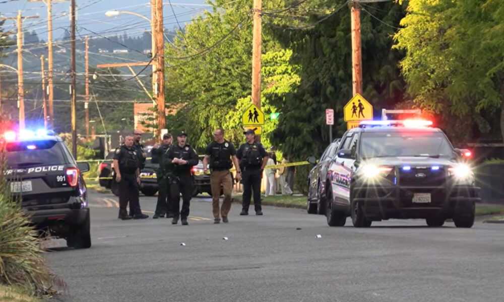 Police officers stand in the middle of a residential street as patrol cars with flashing red and blue lights block the road, with yellow caution tape in the background.