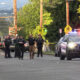 Police officers stand in the middle of a residential street as patrol cars with flashing red and blue lights block the road, with yellow caution tape in the background.