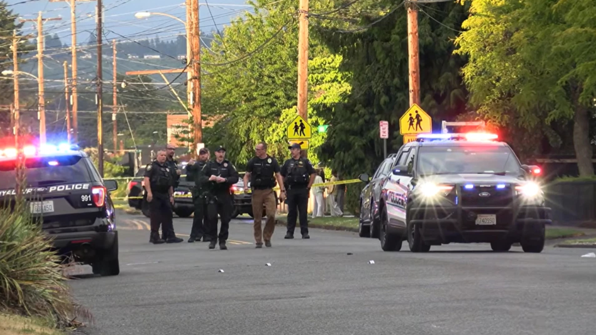Police officers stand in the middle of a residential street as patrol cars with flashing red and blue lights block the road, with yellow caution tape in the background.