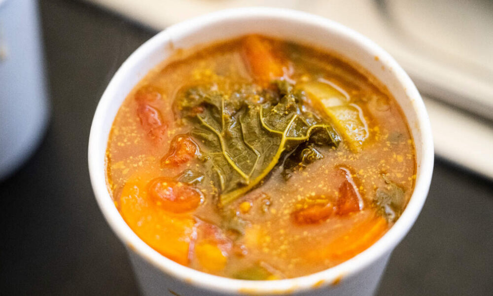 Hot tomato-based vegetable soup in a white cup with a kale leaf on top. Close-up of veggie soup with carrots, tomatoes and a wilted kale leaf. Cup of hearty vegetable soup with leafy greens and orange vegetables. Tomato soup in a cup with visible kale leaf and diced vegetables. Warm bowl of vegetable soup featuring kale and chunky veggies.