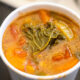 Hot tomato-based vegetable soup in a white cup with a kale leaf on top. Close-up of veggie soup with carrots, tomatoes and a wilted kale leaf. Cup of hearty vegetable soup with leafy greens and orange vegetables. Tomato soup in a cup with visible kale leaf and diced vegetables. Warm bowl of vegetable soup featuring kale and chunky veggies.