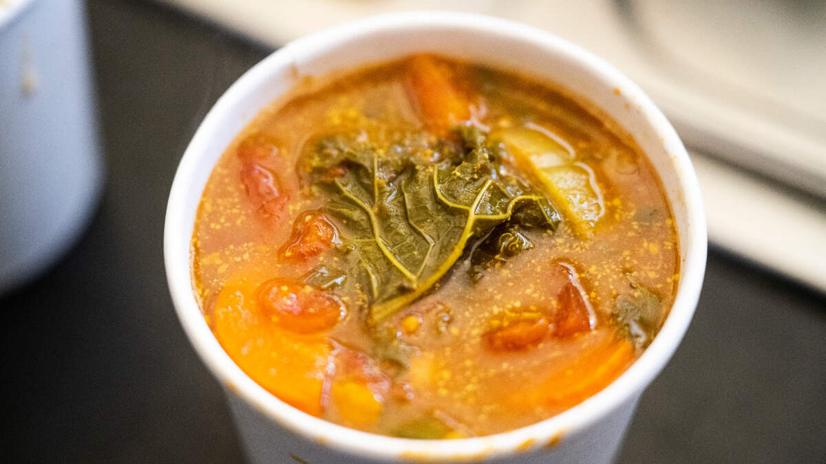 Hot tomato-based vegetable soup in a white cup with a kale leaf on top. Close-up of veggie soup with carrots, tomatoes and a wilted kale leaf. Cup of hearty vegetable soup with leafy greens and orange vegetables. Tomato soup in a cup with visible kale leaf and diced vegetables. Warm bowl of vegetable soup featuring kale and chunky veggies.