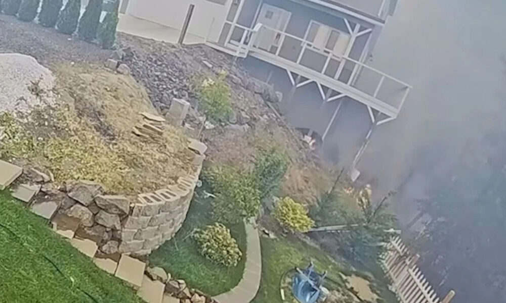 Backyard on a slope featuring a curved stone retaining wall, a stone planter, bushes, and a wooden deck attached to a gray house; smoke or fog covers the right side of the scene.