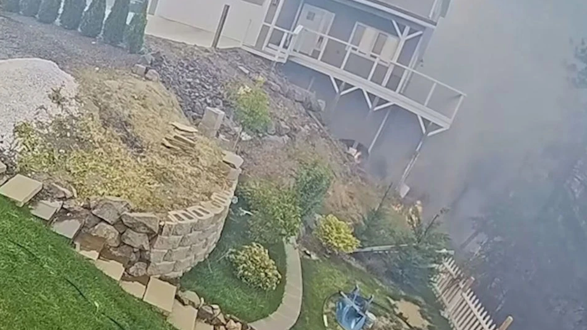 Backyard on a slope featuring a curved stone retaining wall, a stone planter, bushes, and a wooden deck attached to a gray house; smoke or fog covers the right side of the scene.
