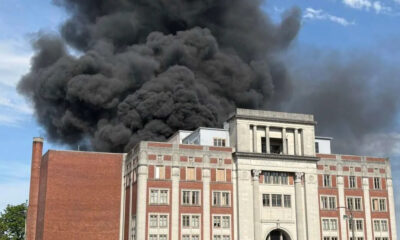 Thick black smoke billowing from the roof of a red-brick campus-style building with a stone entrance. The scene suggests a fire at a public/institutional building on a sunny day.