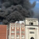 Thick black smoke billowing from the roof of a red-brick campus-style building with a stone entrance. The scene suggests a fire at a public/institutional building on a sunny day.