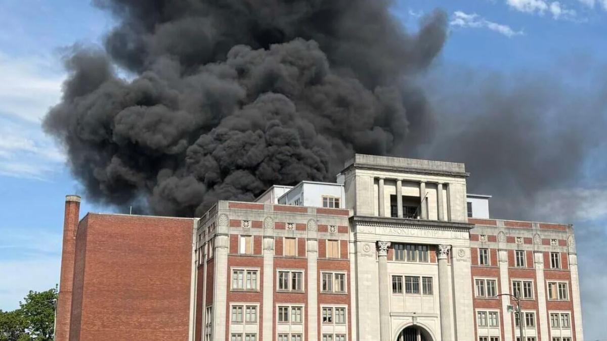 Thick black smoke billowing from the roof of a red-brick campus-style building with a stone entrance. The scene suggests a fire at a public/institutional building on a sunny day.