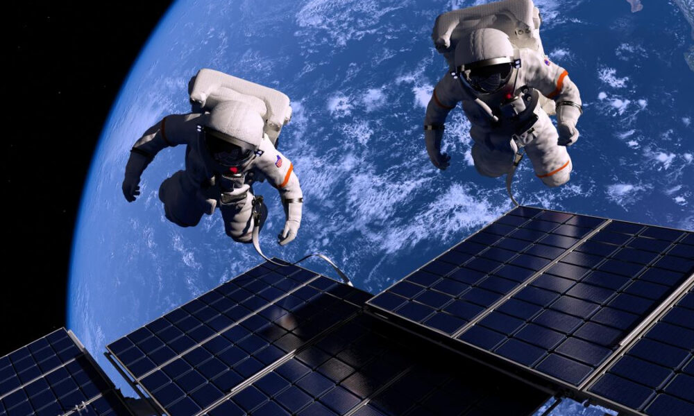 Two astronauts on a spacewalk near solar panels with Earth visible in the background.