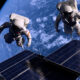 Two astronauts on a spacewalk near solar panels with Earth visible in the background.