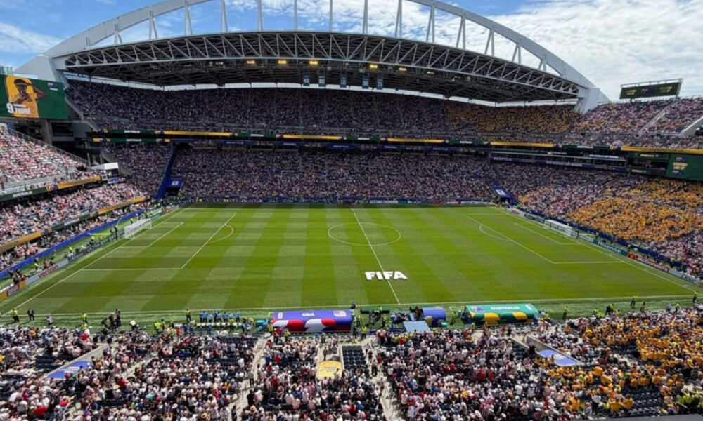Wide-angle view of a full soccer stadium on match day, with the FIFA logo on the grass and fans in every stand under a blue sky.