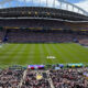 Wide-angle view of a full soccer stadium on match day, with the FIFA logo on the grass and fans in every stand under a blue sky.