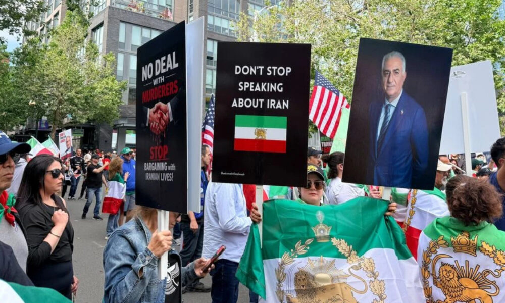 Protesters hold bold signs at a street rally, including 'No deal with murderers' and 'Don’t stop speaking about Iran' with an Iranian flag nearby.