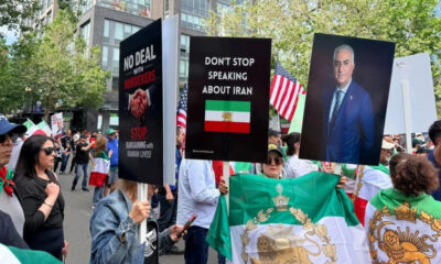 Protesters hold bold signs at a street rally, including 'No deal with murderers' and 'Don’t stop speaking about Iran' with an Iranian flag nearby.