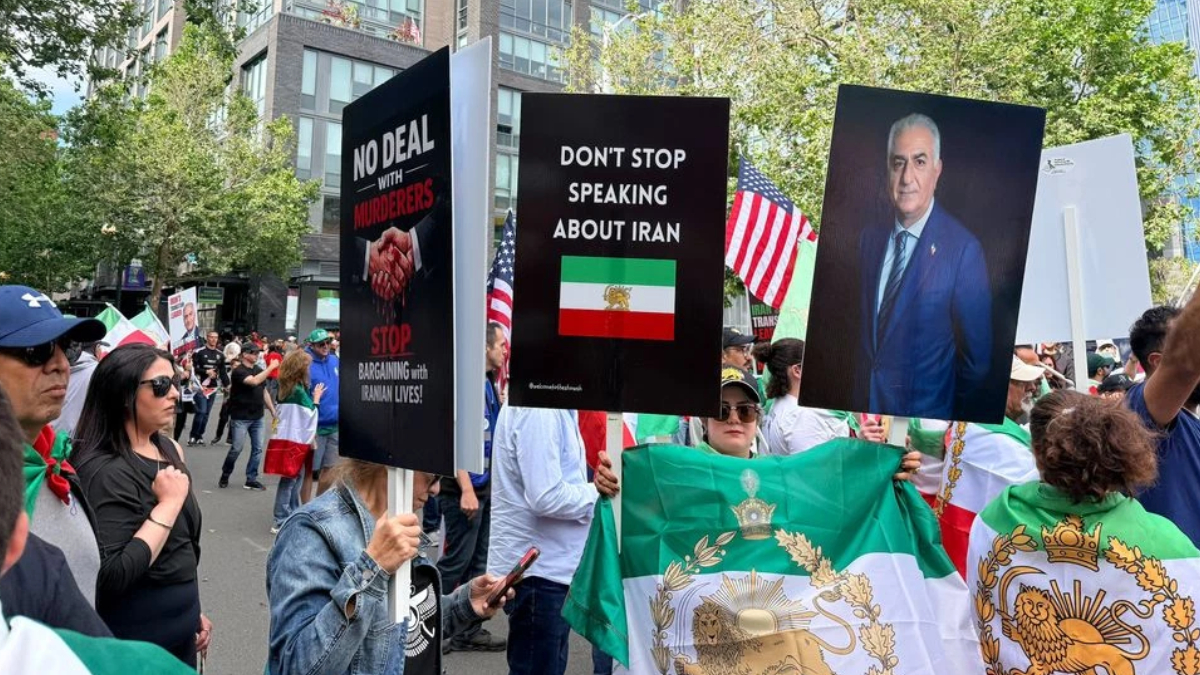 Protesters hold bold signs at a street rally, including 'No deal with murderers' and 'Don’t stop speaking about Iran' with an Iranian flag nearby.