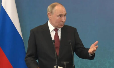 Man in a dark suit giving a speech at a podium with two microphones, Russian flag visible to the left.