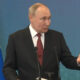 Man in a dark suit giving a speech at a podium with two microphones, Russian flag visible to the left.