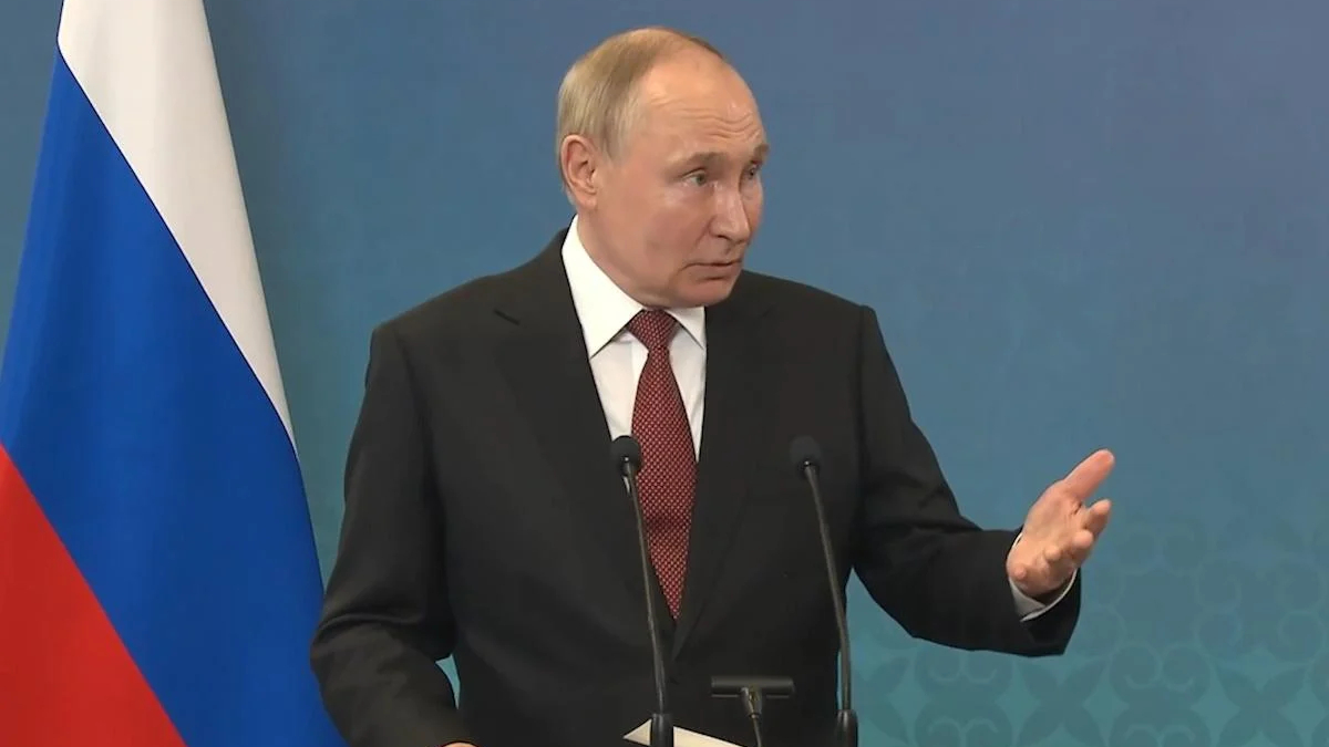 Man in a dark suit giving a speech at a podium with two microphones, Russian flag visible to the left.