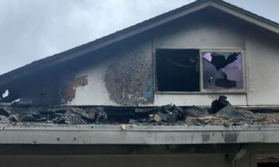 Damaged second-story roof with extensive charring and debris after a fire, a broken window with purple paper inside remains visible under a soot-covered wall.