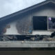 Damaged second-story roof with extensive charring and debris after a fire, a broken window with purple paper inside remains visible under a soot-covered wall.