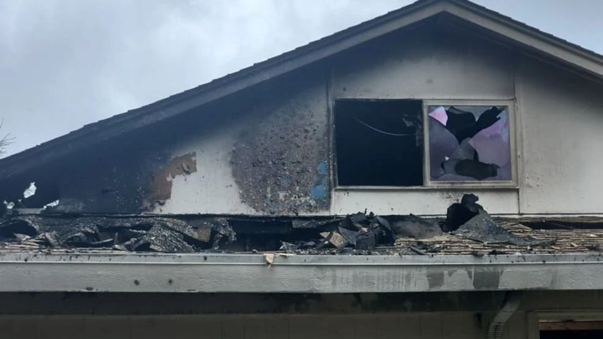 Damaged second-story roof with extensive charring and debris after a fire, a broken window with purple paper inside remains visible under a soot-covered wall.