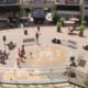 Public square with a circular fountain and people walking around on a sunny day in a mall courtyard.