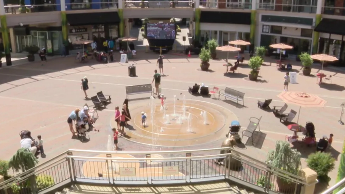 Public square with a circular fountain and people walking around on a sunny day in a mall courtyard.