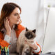 Woman in an orange cardigan working at a desk with a fluffy cat on her lap near a large computer monitor; office workspace in view.
