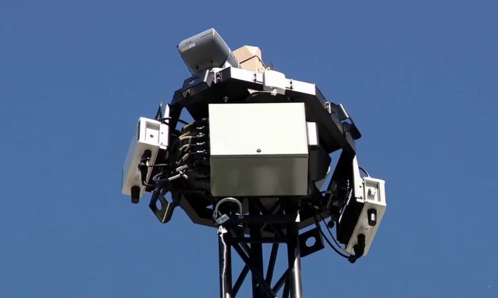 Industrial equipment array mounted on a lattice tower top, with metal enclosures and cables against a clear blue sky