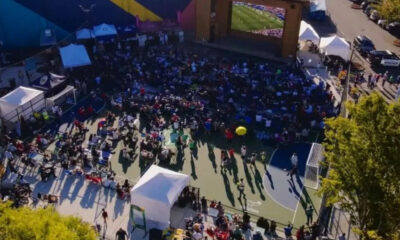Aerial view of an outdoor viewing party on a basketball court, with a large screen stage and a crowd seated and standing around.