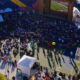 Aerial view of an outdoor viewing party on a basketball court, with a large screen stage and a crowd seated and standing around.