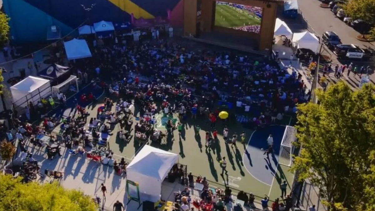 Aerial view of an outdoor viewing party on a basketball court, with a large screen stage and a crowd seated and standing around.
