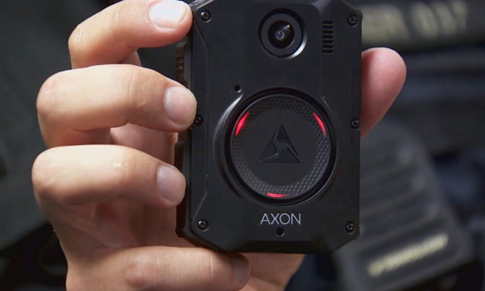 Close-up of a black AXON body camera held in a person's hand, showing the circular lens and red status lights around the edge.