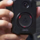Close-up of a black AXON body camera held in a person's hand, showing the circular lens and red status lights around the edge.