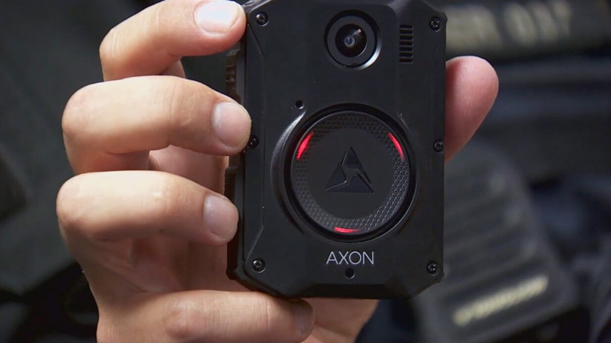 Close-up of a black AXON body camera held in a person's hand, showing the circular lens and red status lights around the edge.
