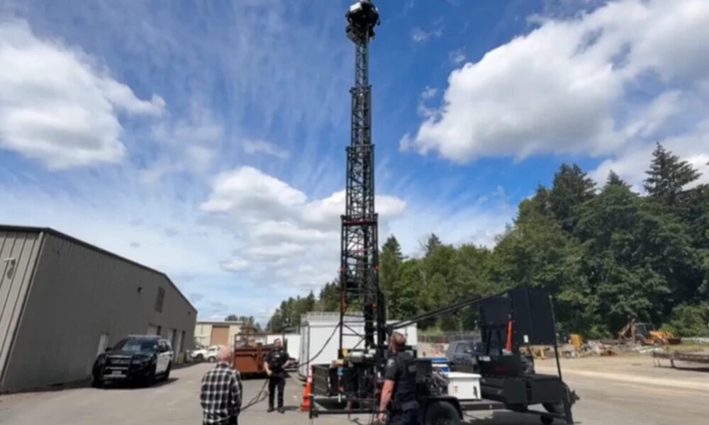 Tall lattice tower on a mobile trailer in an industrial yard, with workers and a pickup truck nearby under a blue sky.