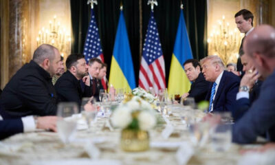 World leaders seated at a long formal table, with Ukrainian and American flags in the background, during a diplomatic meeting.