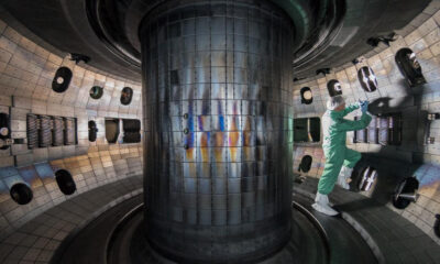 Technician in a cleanroom suit repairin​g equipment inside a large cylindrical vacuum chamber inside a laboratory.