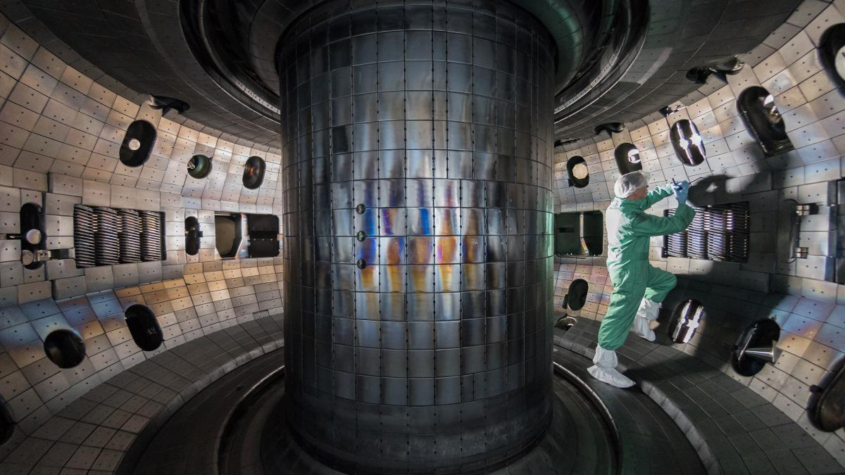 Technician in a cleanroom suit repairin​g equipment inside a large cylindrical vacuum chamber inside a laboratory.
