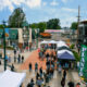 Crowd walking along a brick-paved street lined with vendor tents at a sunny town festival.