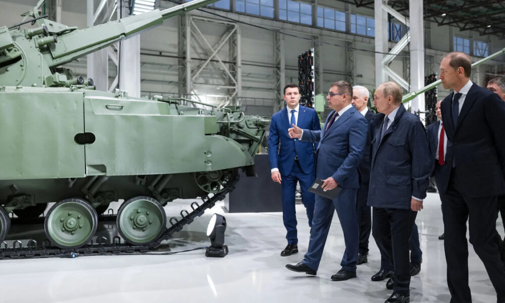 Group of officials in suits examining a large green armored tank on display in a bright hangar-like showroom.