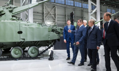 Group of officials in suits examining a large green armored tank on display in a bright hangar-like showroom.
